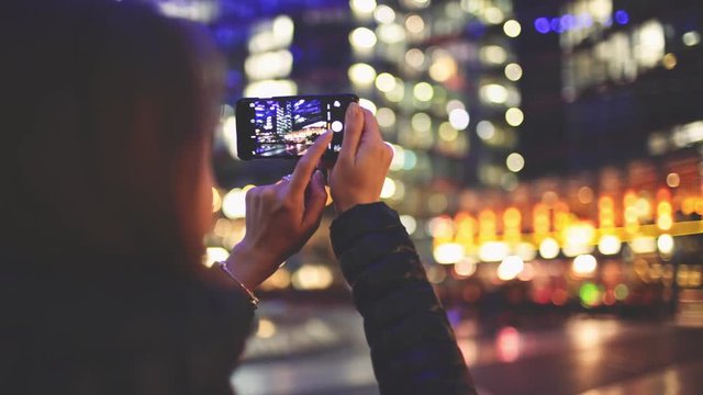 Woman Using SmartPhone at Night in Big City. 4K. Attractive young Woman taking photos on cellphone outdoors. Millennial Girl with phone, blurred Street skyscrapers and traffic lights.
