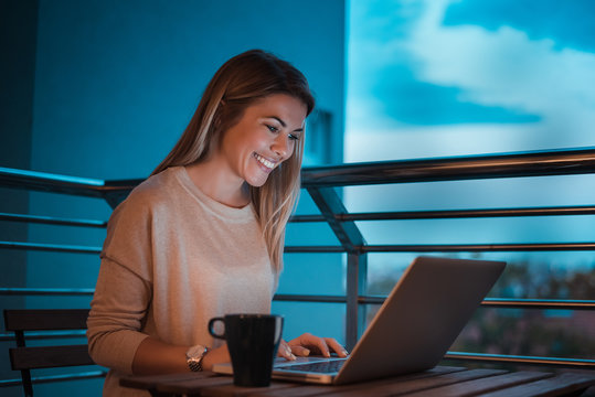 Woman working on a laptop and smiling in balcony at dusk time. High ISO image.