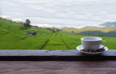 A white cup of hot coffee and coffee beans put interspersed in dish on wood table with green rice field background,wallpaper
