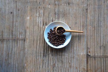 Coffee beans interspersed on white little plate or dish and wooden spoon on bamboo knit wood tray background