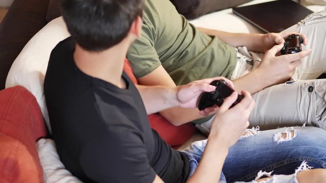 Two young men in casual chilling on sofa and both playing video game console with joysticks in hands