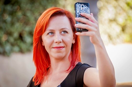 Young and attractive Caucasian girl in her 30s holding the cellular phone in her hands and making a self portrait.