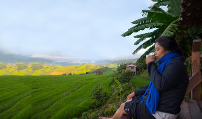 Naklejka premium Woman tourism sitting on wood balcony smelling and drinking hot coffee in morning time and enjoy to see view of green rice field at Napongpeng,Chiangmai-Thailand 