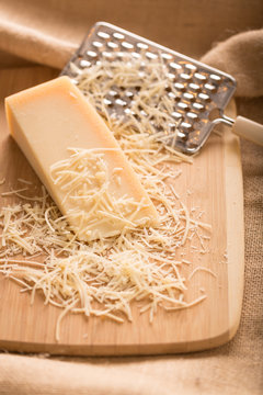 Parmesan Cheese and Grater Medium. an wider view of a block wedge of parmesan cheese with shredded pieces all around and a metal cheese grater on a cutting board