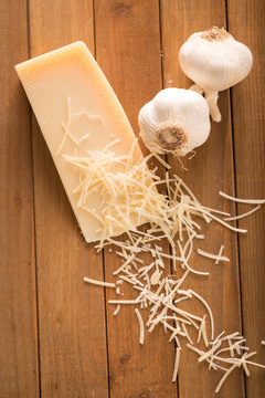 Garlic and Parmesan Cheese Shredded on Wood Board Above. an above shot looking down on a wedge of parmesan cheese and two cloves of garlic with shredded slices of cheese on top