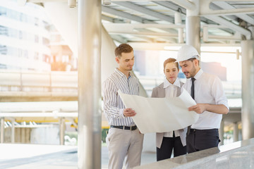 Three people are business manager and engineer meeting project at construction site. Man wearing safety helmet and holding blueprint document.