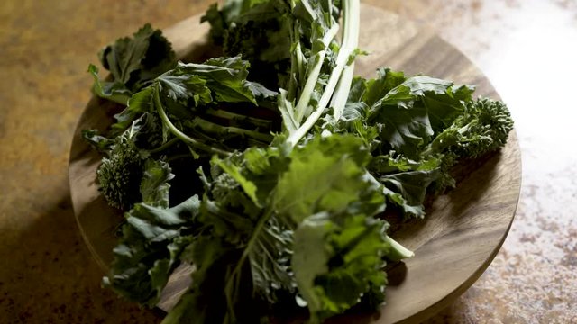 Clusters of stalks of fresh rapini on a round cutting board rotating in beautiful backlit light.