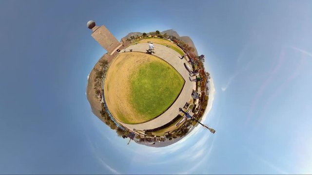 A spherical 360 tiny-planet view of the domed observatory at Ciudad Mitad del Mundo near Quito, Ecuador, a circular pathway, green and dry grass and visitors under a clear blue sky.