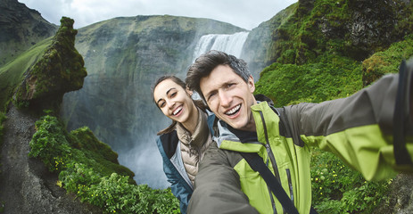 smiling couple, man and woman in warm clothes making selfie on a Skogafoss waterfall background,...