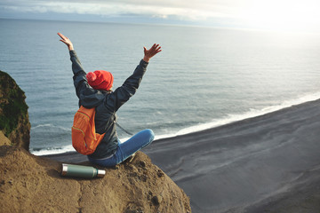 Girl in warm clothing, in red knitted hat and small orange backpack stands on the cliff on background of ocean, sky and Reynisfjara beach in Iceland. Travel to the Iceland