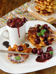 Waffles with cherry and chocolate sauce served on a white plate and in the white gravy boat