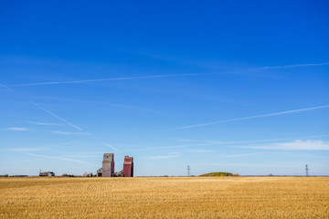 Two old abandoned wooden grain storage elevators with two old buildings beside it in a golden harvested field in a sunny autumn countryside landscape