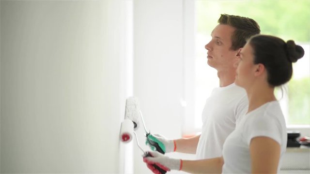 Couple with Platens in Hands Painting the Wall with White Paint in Their Living Room Together. Side View of Woman and Man Doing Apartment Repair.