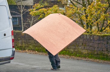 Man with van carries large sheets of wood on his back.