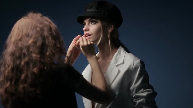 A make-up artist and a stylist work with a model in a black hat