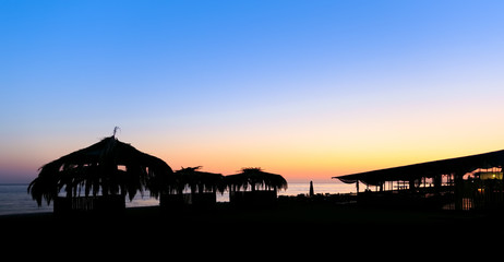 Silhouettes of small hovels with reed roofs and restaurant on the seashore at sunset