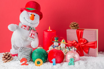 Christmas or New Year card. Funny snowman with burning candle, cones, giftbox, balls and 2018 figures on snow against red background. Focus on figures.