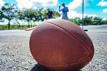close up of an american football on the ground with people playing cornhole in the background while tailgating before the game