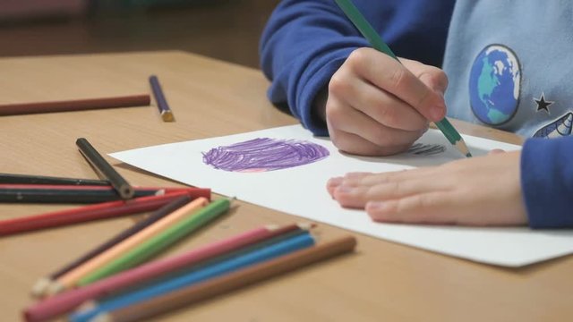 Close-up of hands of unidentified little boy in blue sweater sitting at table drawing pictures using multicolored pencils in kindergarten