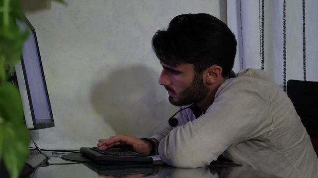 Young male home worker with headset and microphone falling asleep as he's working from his living room in front of computer late at night