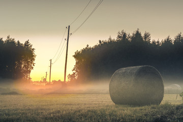  Sunset in countryside at mid summer. Hay rolls and road in misty weather with clear sunny sky....