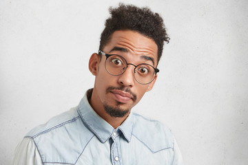 Portrait of young Afro American man with bugged eyes, has trendy hairdo, mustache and beard, looks puzzled as finds out unexpected facts or information, isolated over white studio background