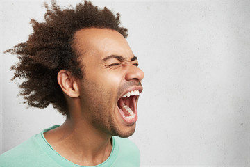 Horizontal portrait of man with dark skin and Afro hairstyle screams in despair, opens mouth...