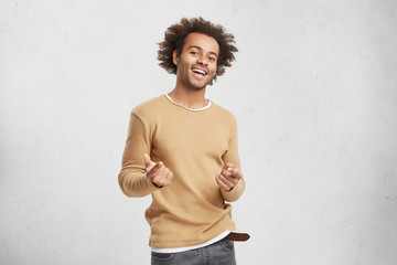 Stylish happy African American male wears casual clothes, points in camera with fore fingers,...