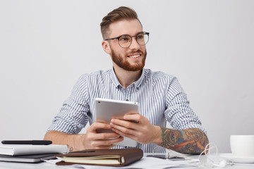 Portrait of successful office worker in round spectacles, has tattooed arms, holds modern tablet, makes annual report, going to have minute rest and surf social networks. Technolgy, business concept