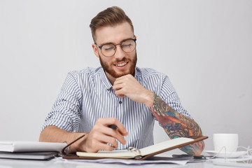 Portrait of stylish hipster guy with colored tattoo on hand, writes notes in notebook, drinks coffee, sits at office. Young male student prepare report, puts down main ideas in diary not to forget