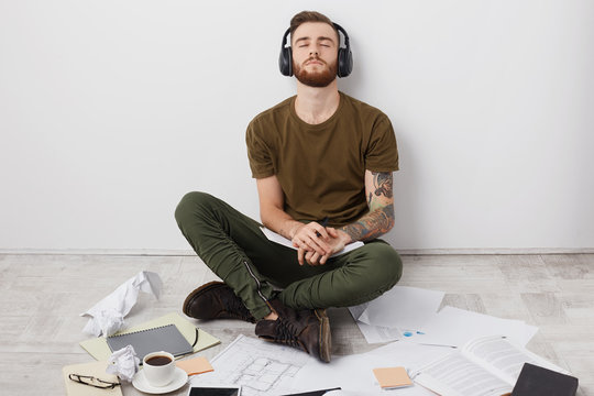 Relaxed hipster student in casual outfit, sits on floor crossed legs, enjoys rock music in headphones, closes eyes with delight, surrounded with papers and books, prepares for seminar in literature