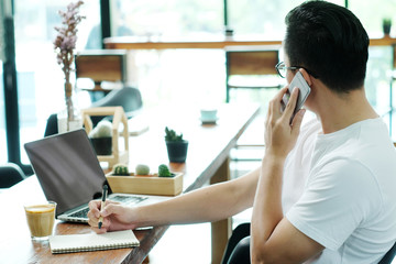 Young asian man talking phone while writing on notebook paper and working on laptop computer at cafe table background, business casual lifestyle, online education
