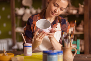 A dark-haired young woman, a potter in a plaid shirt and apron, paints a clay cup in a gray color on a wooden table, on which there are tools, and in the background in the workshop clay vases