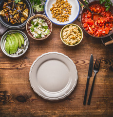 Tasty vegetarian ingredients for healthy meal in bowls: chick peas puree, roasted vegetables , red paprika stew, avocado and seeds served with empty plate with cutlery Clean eating food concept