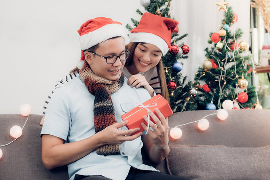 Asia lovers couple,girlfriend surprise boyfriend by giving Christmas present at sofa with xmas decoration tree at house party,Holiday celebrating season.