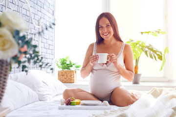 Obraz na płótnie Canvas Pregnant woman drinking a cup of tea with macaroons on bed