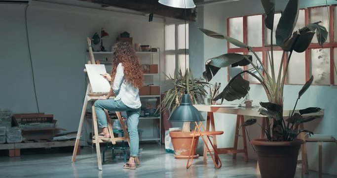 Beautiful long-haired woman draws a sketch on a blank canvas in her large studio