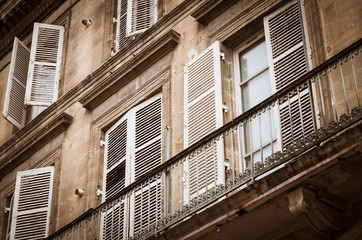 Old windows with weathered shutters in a facade of a kind of mansion with a balcony with forged ornamental fence, viewing point is oblique which. Coloring is somewhat bleak and gray.