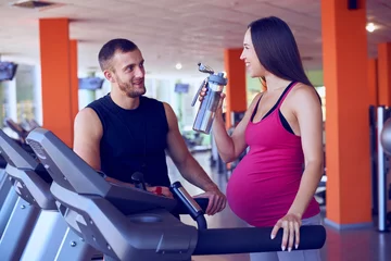 Gordijnen Pregnant woman drinking water while speaking to personal trainer in gym © ArtFamily