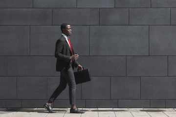 Full-length portrait of handsome African American entrepreneur walking on pavement with gray block wall in background, dressed in black suit, holding leather bag and coffee cup during break in work