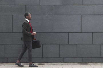 Horizontal shot of young dark-skinned businessman spending time from work walking in city center in afternoon time, drinking coffee and carrying business bag while moving from one meeting to another