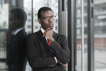 Horizontal picture of young African American male dressed in formal clothes standing in his office, looking through window, scratching chin as if thinking of new strategies for promotion of business