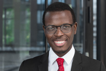 Horizontal headshot of handsome dark-skinned businessman with glass background behind, spending time in office during break with spectacles on, smiling friendly as sign of trust and openness