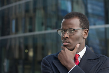 Horizontal headshot of attentive African american executive standing outdoors, casting side look through eyeglasses scratching chin with fingers, thinking
