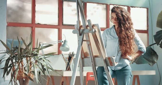 Portrait of a beautiful long-haired female artist at work at her canvas in a studio