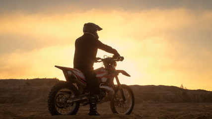 Professional FMX Motorcycle Rider Rests on His Bike and Overlooks Hard Sandy Off-Road Terrain. Sun...