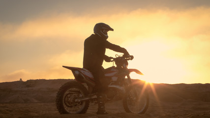 Fototapeta na wymiar Professional FMX Motorcycle Rider Rests on His Bike and Overlooks Hard Sandy Off-Road Terrain. Sun is Setting.