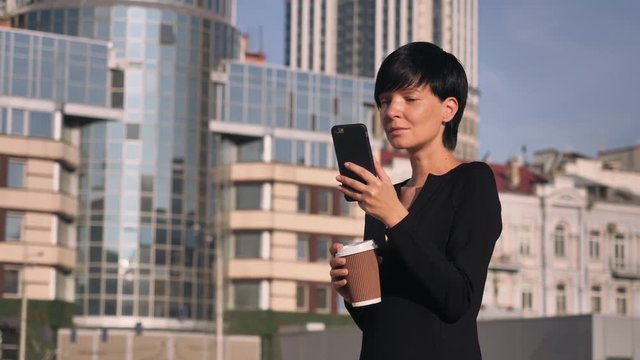 Young brunette scrolling touch screen smartphone outdoors. Happy businesswoman spend coffee break outdoors. Smiling woman standing near office building texting message on smart phone. Caucasian lady