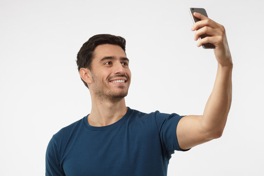 Portrait of young handsome caucasian man standing against gray background using smartphone to take selfie pictures and smiling