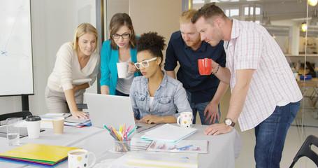 Young people standing around female coworker and watching laptop at table in office all together sharing ideas.